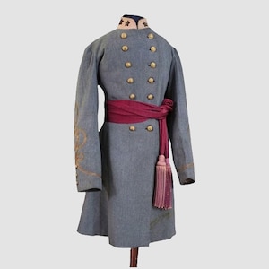 Könnte beinhalten: Ein grauer Militärmantel mit goldenen Knöpfen und Verzierungen, einem hohen Kragen und einer weinroten Schärpe mit Quasten. Der Mantel hat goldene Stickereien an den Ärmeln und wird auf einem Ständer ausgestellt. Der Mantel scheint Vintage oder historisch zu sein.