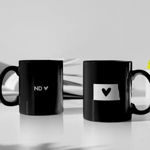 May include: Two black coffee mugs. One mug features the letters "ND" with a heart symbol, and the other displays a white outline of the state of North Dakota with a heart. The mugs are on a white surface.