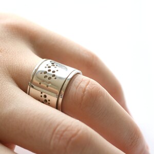 May include: A silver ring with a wide band featuring a series of small, irregularly shaped cut-out holes. The ring is worn on a finger, and the design is visible against the skin. The ring appears to be made of polished metal.