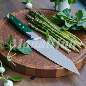 Könnte beinhalten: Ein Edelstahl-Kochmesser mit grünem Griff liegt auf einem Holzschneidebrett neben frischem Spargel. Das Messer hat eine scharfe Klinge und einen genieteten Griff. Das Schneidebrett ist rund mit dunkler und hellbrauner Holzmaserung.