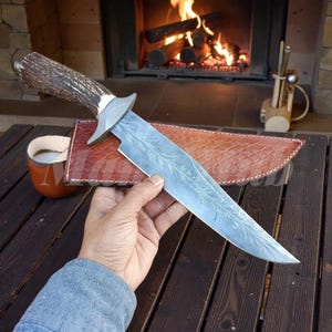 Puede incluir: Un cuchillo de caza grande con una hoja grabada decorativa, mango de asta y una funda de cuero. El cuchillo se sostiene sobre una mesa de madera, con una chimenea y una taza en el fondo.