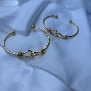 Puede incluir: Dos brazaletes dorados de extremo abierto con un diseño de nudo en el centro. Cada brazalete tiene una pequeña bola en cada extremo. Los brazaletes se exhiben sobre una tela blanca y sedosa.