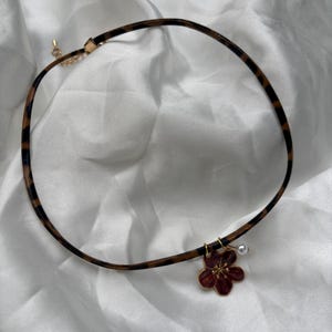 May include: A choker necklace with a brown and black leopard print band. A dark red flower pendant with gold accents and a small white pearl hangs from the band. The necklace is on a white background.