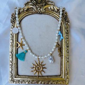 May include: A pearl necklace with gold and turquoise charms, including a starfish, tassel, seahorse, and sun pendant. The necklace is displayed on a gold ornate frame.
