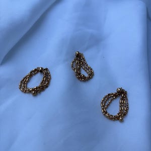 Peut inclure: Trois bagues en chaîne dorée, chacune avec un design unique, reposent sur un tissu blanc. Les bagues sont composées de plusieurs brins de chaînes délicates, créant un aspect texturé. L'éclairage met en valeur l'éclat métallique des bijoux.