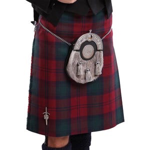 May include: A red and green tartan kilt featuring a silver kilt pin with a sword design. A grey fur sporran with a silver metal top and three hanging tassels is attached to the waist by a silver chain. The kilt is worn with a black jacket.