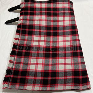 May include: A kilt featuring a red, black, and white tartan plaid pattern. The garment includes two black leather straps with metal buckles on the side. The edge of the fabric has a frayed, unfinished fringe detail. The text Scotland Craft is visible.
