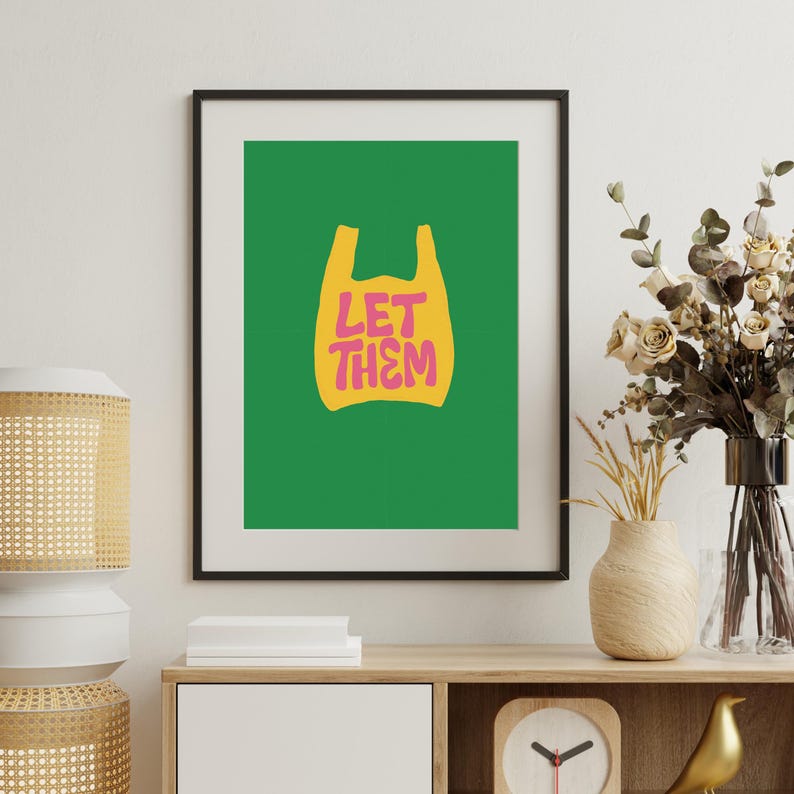 May include: Framed art print with a green backdrop and a yellow shopping bag graphic. The bag displays the pink words "LET THEM". The artwork is displayed on a wooden cabinet with decorative items.