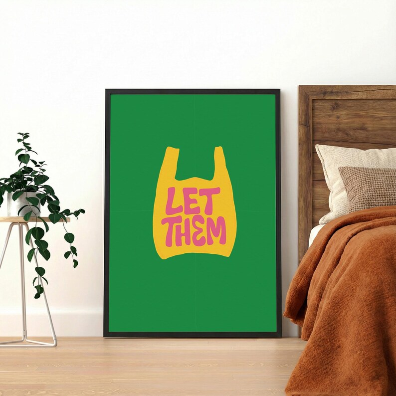 May include: A framed art print with a green background and a yellow shopping bag shape. The words "LET THEM" are written in pink within the bag shape. The print is displayed on a wooden floor, with a plant and bed visible.