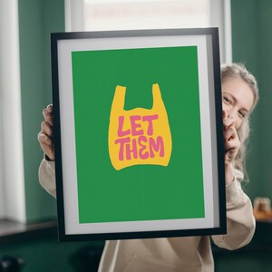 May include: A framed art print with a green background and a yellow shopping bag graphic. The words "LET THEM" are printed in pink within the bag. The frame is black.
