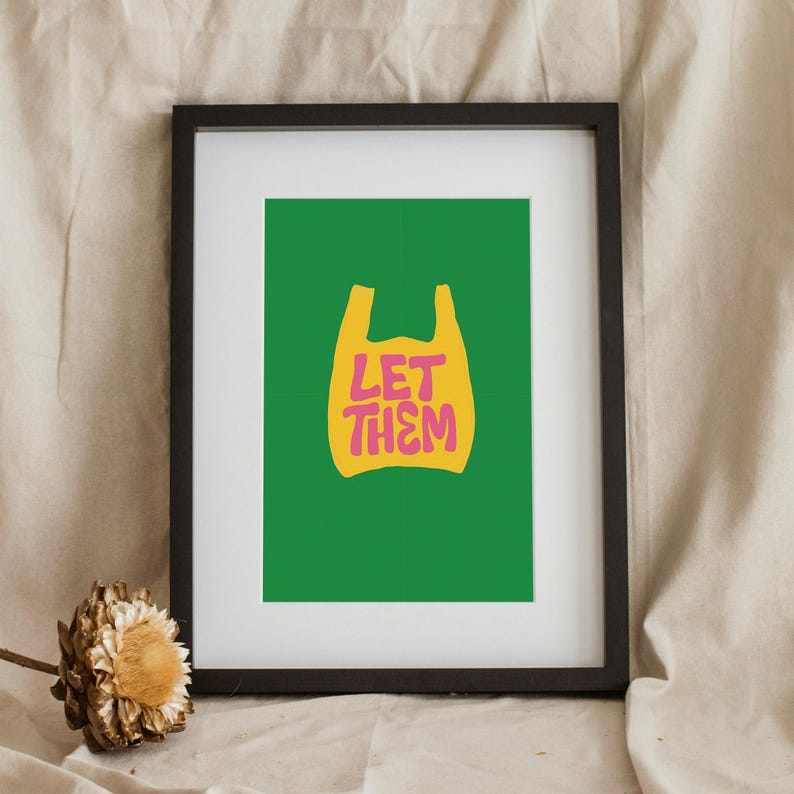 May include: Framed art print with a green background and a yellow shopping bag graphic. The pink text within the bag reads "LET THEM". The black frame surrounds the print. A dried flower is in the lower left corner.