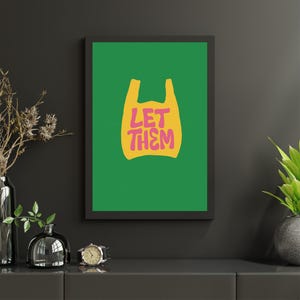 May include: A framed art print with a green background and a yellow plastic bag graphic. The pink text "LET THEM" is inside the bag shape. The artwork is mounted on a dark grey wall.