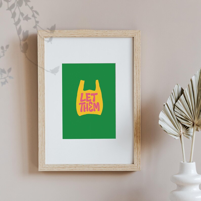 May include: Framed art print with a green backdrop and a yellow plastic bag graphic. The pink text "LET THEM" is inside the bag shape. The frame is made of light-coloured wood.