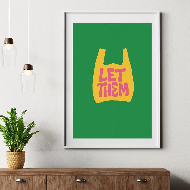 May include: A framed art print with a yellow plastic bag graphic and the pink text "LET THEM". The background is green. The print is in a white frame, displayed on a wooden surface.