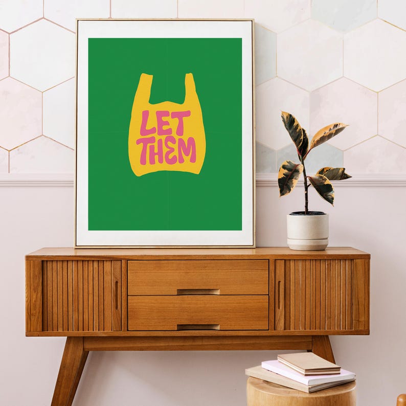 May include: A framed art print with a yellow shopping bag design and the pink text "LET THEM" on a green backdrop. The print is displayed on a wooden cabinet. A plant in a white pot is also visible.