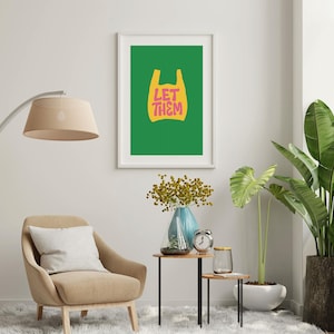 May include: A framed print featuring a green background with a yellow shopping bag graphic and the pink text "LET THEM". The artwork is displayed in a living room with a beige armchair, a floor lamp, and potted plants.