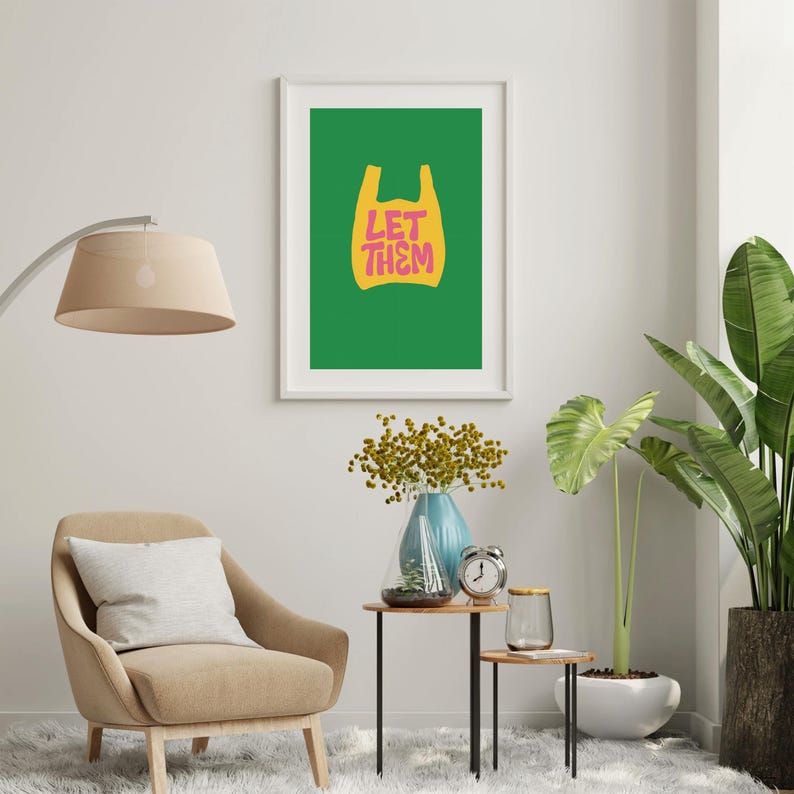 May include: A framed print featuring a green background with a yellow shopping bag graphic and the pink text "LET THEM". The artwork is displayed in a living room with a beige armchair, a floor lamp, and potted plants.