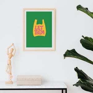 May include: A framed art print featuring a green background and a yellow shopping bag graphic. The words "LET THEM" are in pink and yellow. The print is in a light wood frame and is displayed on a white wall.