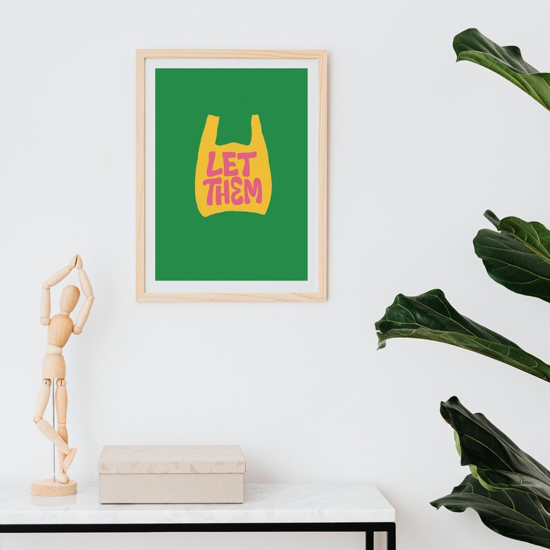 May include: A framed art print featuring a green background and a yellow shopping bag graphic. The words "LET THEM" are in pink and yellow. The print is in a light wood frame and is displayed on a white wall.