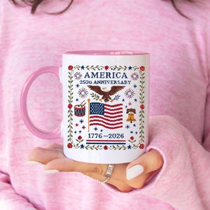 Vintage Americana cross-stitch mug styled on a wooden table with eucalyptus, white ceramic with patriotic eagle and US flag art celebrating America's 250th birthday.