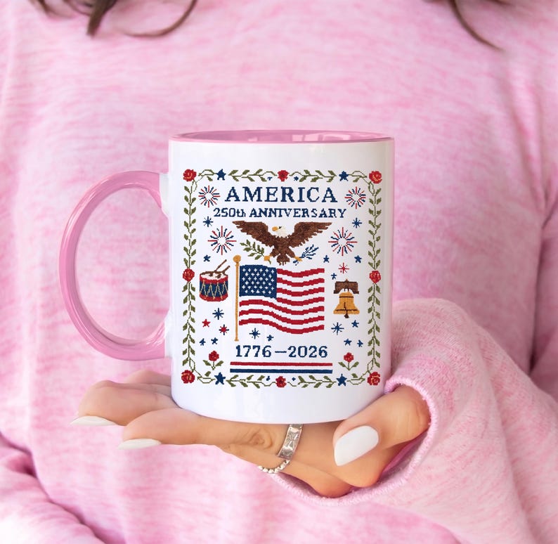 Vintage Americana cross-stitch mug styled on a wooden table with eucalyptus, white ceramic with patriotic eagle and US flag art celebrating America's 250th birthday.