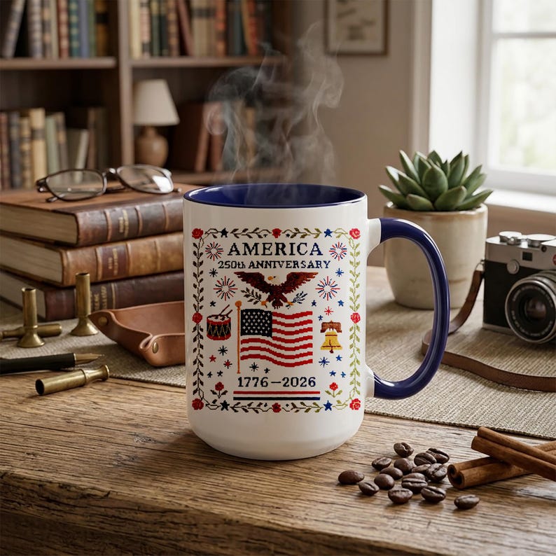 Side angle of the 250th anniversary patriotic mug with handle visible, white ceramic printed with folk-art cross-stitch American flag and eagle for the 2026 semiquincentennial.