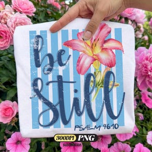 Pode incluir: Camiseta branca com listras verticais azuis e as palavras "be still" em fonte cursiva. Uma flor de lírio rosa e amarela em estilo aquarela está na camiseta. O texto "Psalm 46:10" está abaixo. A camiseta é segurada em frente a flores cor-de-rosa.