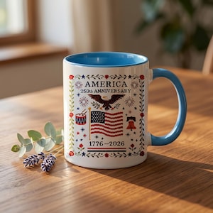 Hand holding the America 250th anniversary cross-stitch mug, showing scale and the red-white-and-blue eagle, flag and Liberty Bell sampler design dated 1776–2026.