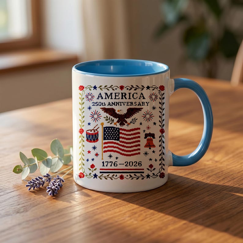 Hand holding the America 250th anniversary cross-stitch mug, showing scale and the red-white-and-blue eagle, flag and Liberty Bell sampler design dated 1776–2026.