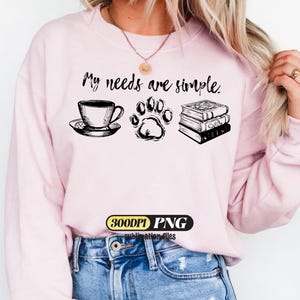 May include: Pink sweatshirt with the text "My needs are simple." The design includes a teacup and saucer, a paw print, and a stack of books, all in black ink. The image also includes a "300DPI PNG" logo.