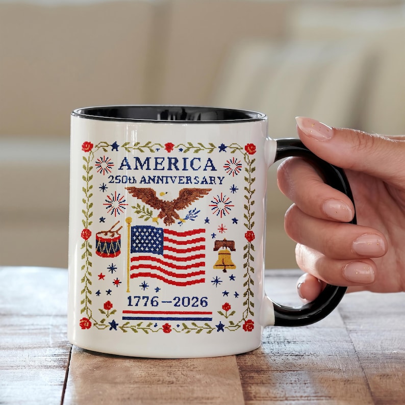 White ceramic mug with cross-stitch–style design of an American eagle, US flag, Liberty Bell and drum, reading "America 250th Anniversary 1776–2026" — vintage Americana semiquincentennial patriotic gift.