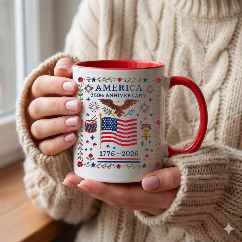 Close-up of the cross-stitch patriotic design on a white coffee mug, showing the embroidered bald eagle, fireworks and rose-vine border of the America 250th anniversary 1776–2026 keepsake.