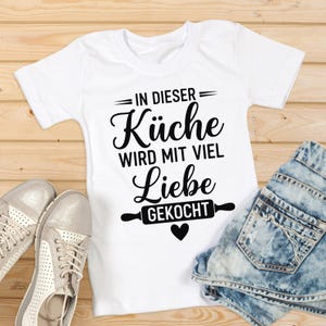May include: White t-shirt with the German text "IN DIESER K&uuml;che WIRD MIT VIEL Liebe GEKOCHT" in black script, featuring a rolling pin graphic and a heart. The shirt is paired with denim shorts and silver sneakers.