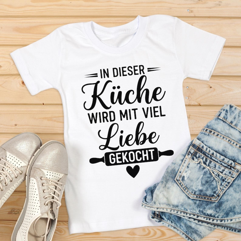 May include: White t-shirt with the German text "IN DIESER K&uuml;che WIRD MIT VIEL Liebe GEKOCHT" in black script, featuring a rolling pin graphic and a heart. The shirt is paired with denim shorts and silver sneakers.