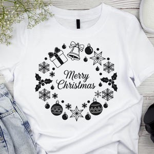 Op de afbeelding: Wit T-shirt met een zwart-wit kerstkransontwerp. De krans bevat een geschenkdoos, bel, ballen, sneeuwvlokken, sterren en hulst. De tekst "Merry Christmas" staat in het midden.