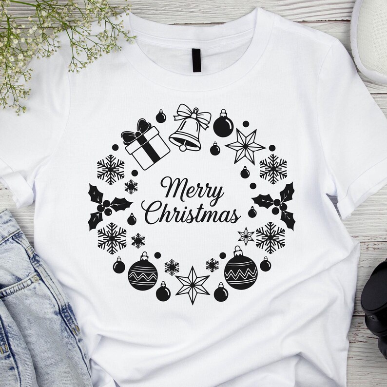 Op de afbeelding: Wit T-shirt met een zwart-wit kerstkransontwerp. De krans bevat een geschenkdoos, bel, ballen, sneeuwvlokken, sterren en hulst. De tekst "Merry Christmas" staat in het midden.