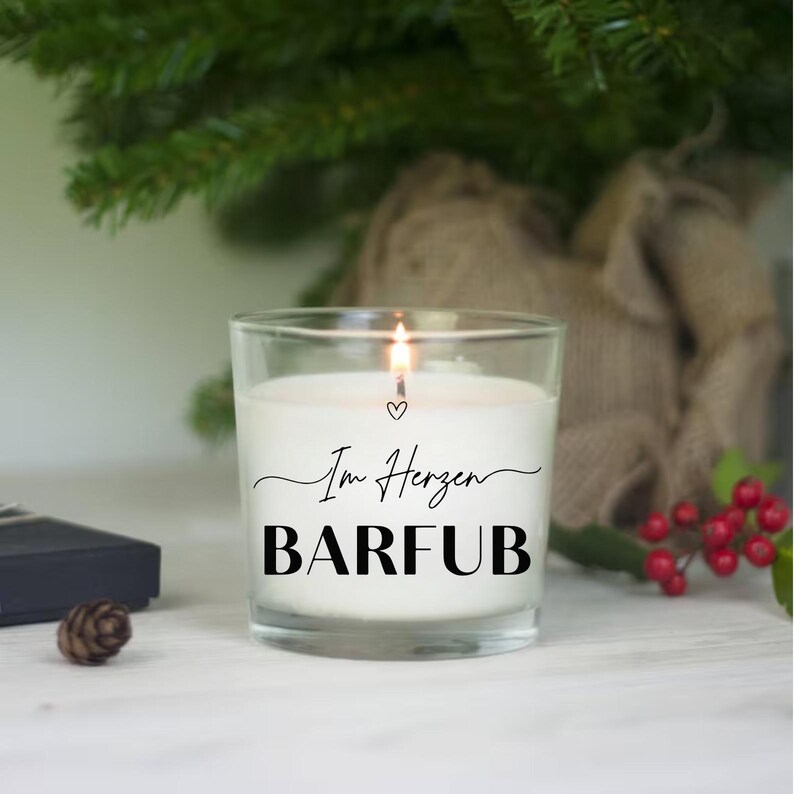 Puede incluir: Una vela blanca encendida en un tarro de cristal transparente. La vela tiene el texto negro "Im Herzen BARFUB" con un s&iacute;mbolo de coraz&oacute;n. El fondo incluye un &aacute;rbol de Navidad y bayas rojas.
