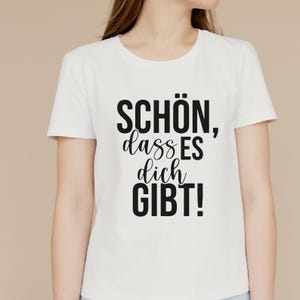 Op de afbeelding: Wit T-shirt met de Duitse tekst "SCH&Ouml;N, dass ES dich GIBT!" in zwart, een positieve bevestiging. Het shirt heeft een ronde hals en korte mouwen. De tekst staat in het midden op de voorkant.