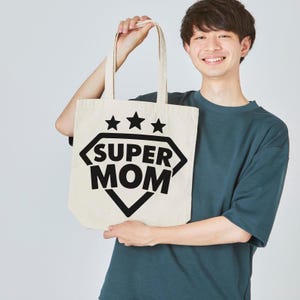 Op de afbeelding: Een cr&egrave;mekleurige draagtas met de tekst "SUPER MOM" in een zwarte ruitvorm, met drie zwarte sterren erboven. De tas wordt door een persoon vastgehouden.