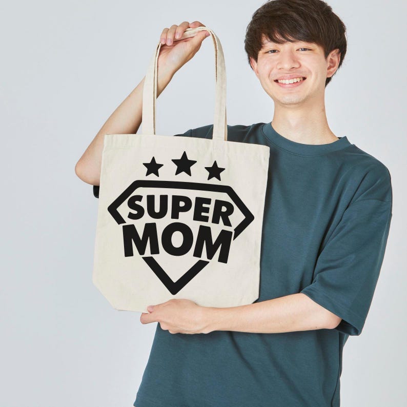 Op de afbeelding: Een cr&egrave;mekleurige draagtas met de tekst "SUPER MOM" in een zwarte ruitvorm, met drie zwarte sterren erboven. De tas wordt door een persoon vastgehouden.