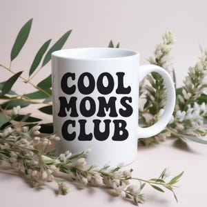 Op de afbeelding: Een witte keramische mok met de tekst "COOL MOMS CLUB" in vette, zwarte letters. De mok heeft een gebogen handvat en is omgeven door groene en witte bloemstukken tegen een zachtroze achtergrond.