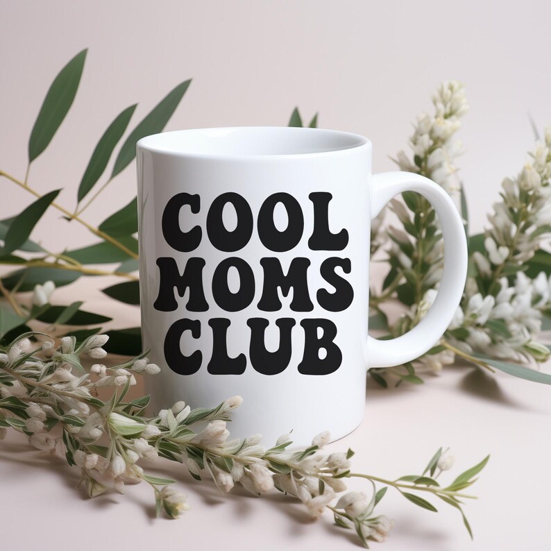 Op de afbeelding: Een witte keramische mok met de tekst "COOL MOMS CLUB" in vette, zwarte letters. De mok heeft een gebogen handvat en is omgeven door groene en witte bloemstukken tegen een zachtroze achtergrond.