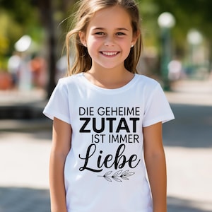 May include: A white t-shirt with the German text "DIE GEHEIME ZUTAT IST IMMER Liebe" in black. The word "Liebe" is in a cursive font with a sprig of leaves below it. The shirt is being worn by a young girl.