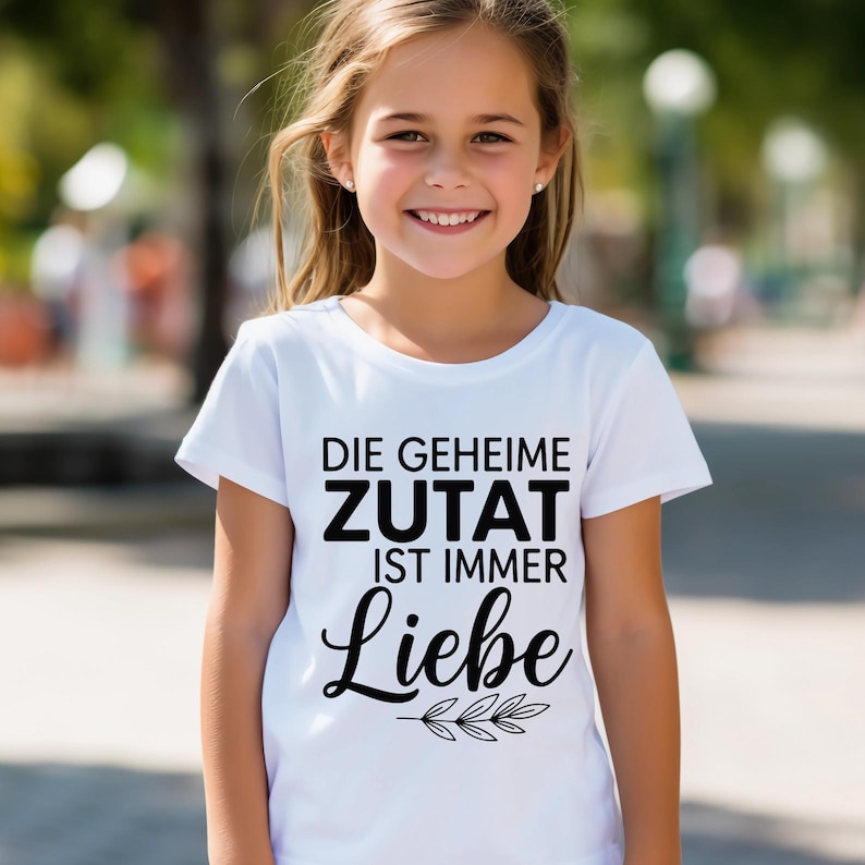 May include: A white t-shirt with the German text "DIE GEHEIME ZUTAT IST IMMER Liebe" in black. The word "Liebe" is in a cursive font with a sprig of leaves below it. The shirt is being worn by a young girl.