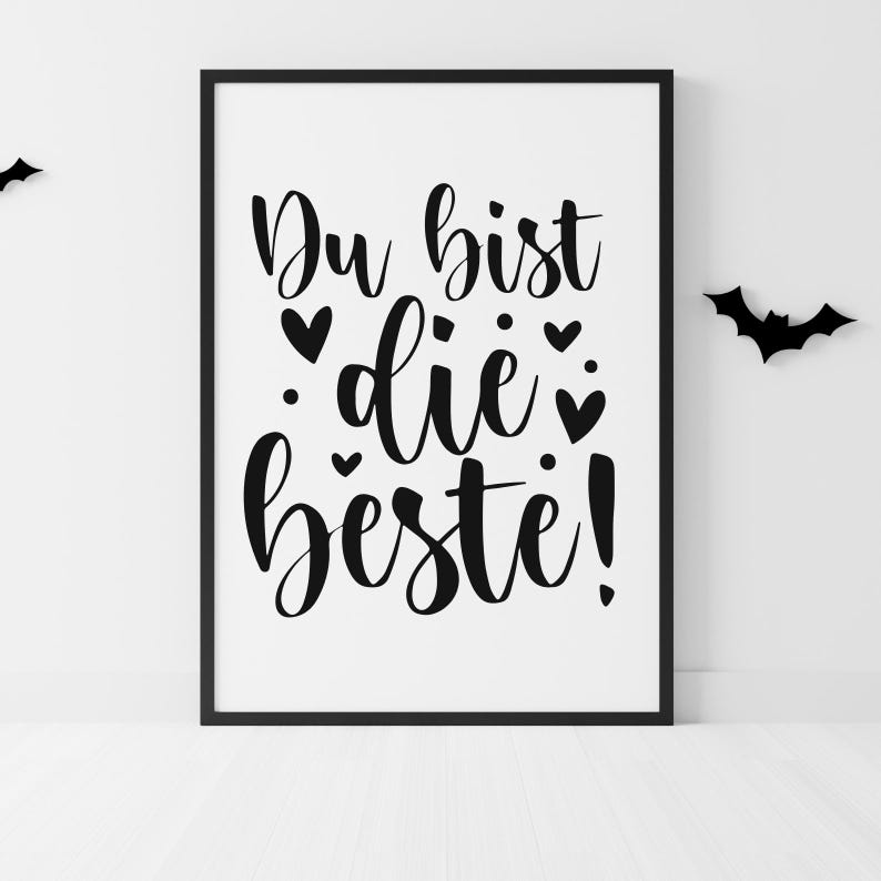 Op de afbeelding: Een ingelijste print met de Duitse zin "Du bist die Beste!" in zwart schrift, wat "Je bent de beste!" betekent. De witte achtergrond is versierd met zwarte harten en stippen. Twee zwarte vleermuis silhouetten staan op de muur.