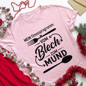 May include: A light pink t-shirt with the German text "MEIN fitnessprogramm: VOM Blech ZUM MUND" in black. The design includes a fork, spoon, and arrows. The shirt is a crew neck with rolled-up sleeves.