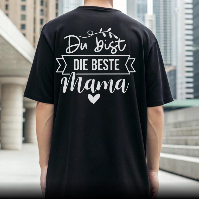 Op de afbeelding: Zwart T-shirt met witte tekst "Du bist DIE BESTE Mama" en een hartsymbool. De tekst is in een decoratieve stijl, met "Die Beste" in een banner. Het T-shirt is een casual stijl.