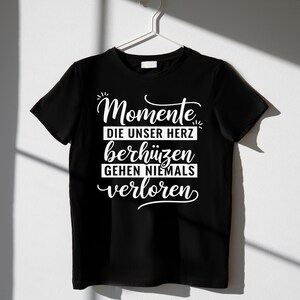 Puede incluir: Camiseta negra con la frase alemana "Momente die unser Herz ber&uuml;hren gehen niemals verloren" en escritura blanca. La camiseta est&aacute; colgada de una percha blanca sobre un fondo blanco. El texto se traduce como "Los momentos que tocan nuestros corazones nunca se pierden."