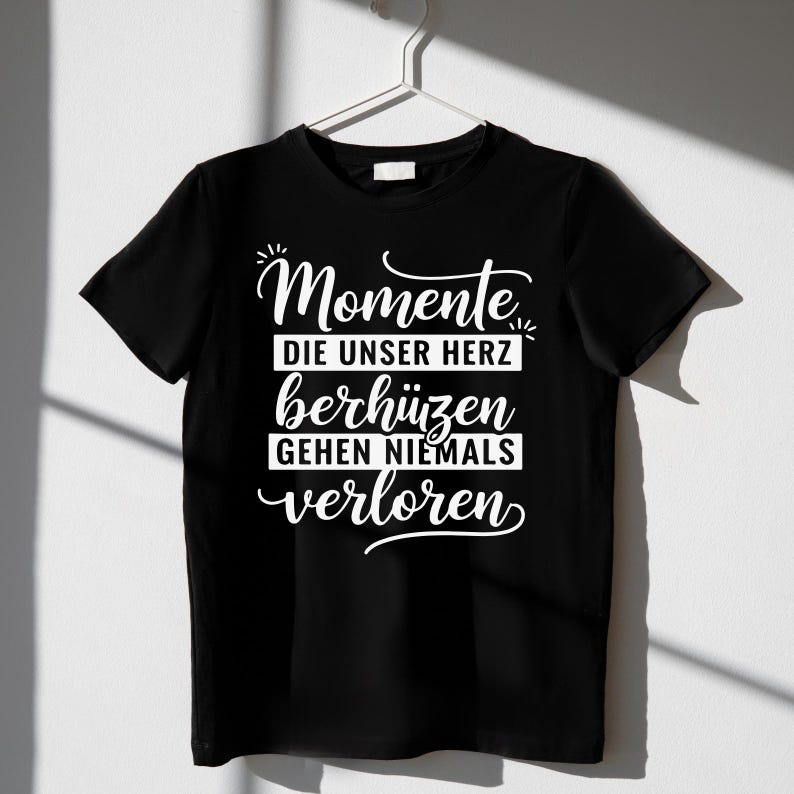 Puede incluir: Camiseta negra con la frase alemana "Momente die unser Herz ber&uuml;hren gehen niemals verloren" en escritura blanca. La camiseta est&aacute; colgada de una percha blanca sobre un fondo blanco. El texto se traduce como "Los momentos que tocan nuestros corazones nunca se pierden."
