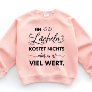 Puede incluir: Una sudadera rosa claro con el texto en alem&aacute;n "EIN L&auml;cheln KOSTET NICHTS aber es ist VIEL WERT" en negro. El texto incluye dos s&iacute;mbolos de coraz&oacute;n y escritura decorativa.